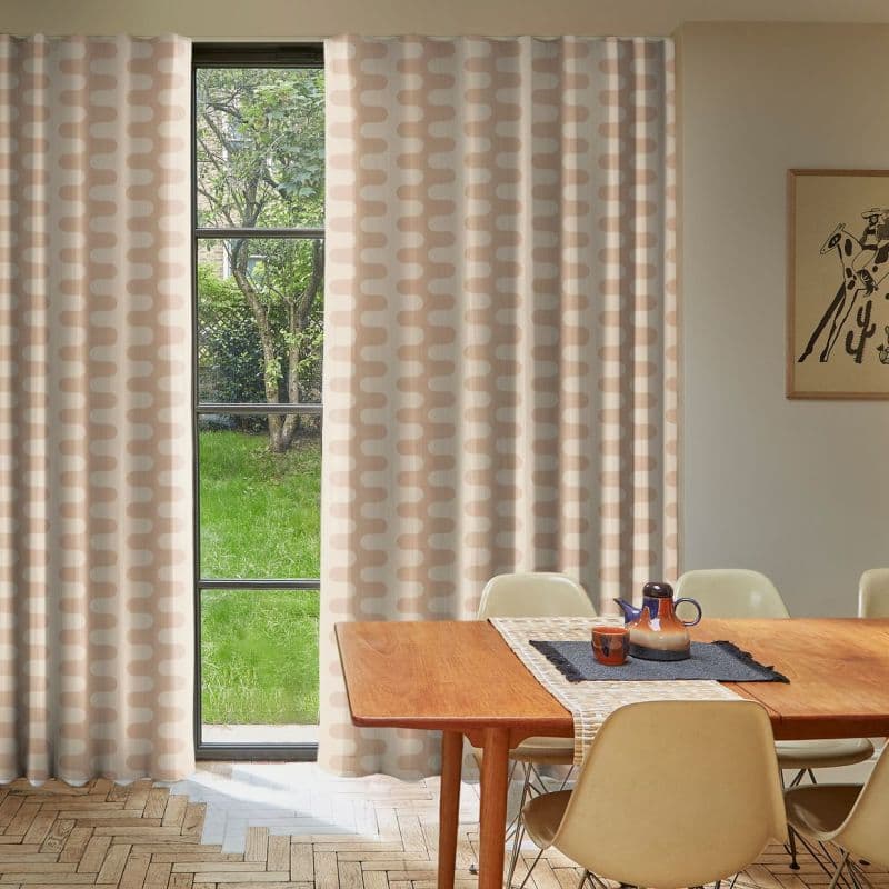Beige-and-white patterned floor-length curtains, hung beside a tall black-framed window, are parted to reveal a green yard; wooden dining table with chairs sits in a sunlit, minimalist dining room.