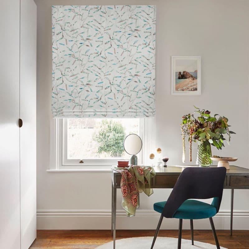 Patterned Roman blind, lowered over sash window with pale leaf motif and soft folds, filtering daylight; above a desk with chair, mirror and vase of flowers in a bright room.