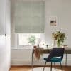 Fabric Roman blind with pale green circular motif, lowered over the window and filtering soft daylight through its folds; a desk, chair, mirror and vase of foliage in an office.