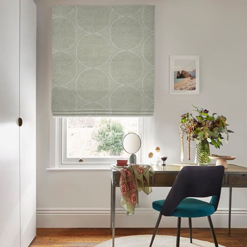 Fabric Roman blind with pale green circular motif, lowered over the window and filtering soft daylight through its folds; a desk, chair, mirror and vase of foliage in an office.
