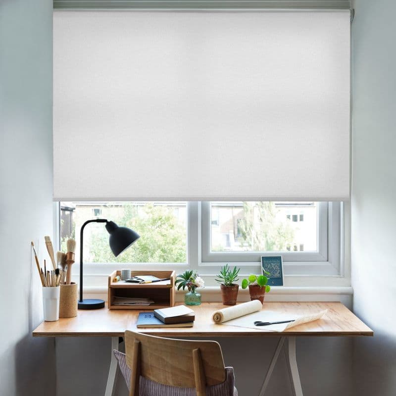 White fabric roller blind, lowered over the window with a straight hem, filtering soft daylight. Below, a wooden desk workspace with lamp, brushes, notebooks, small potted plants and suburban view.