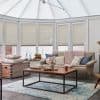 Cellular honeycomb pleated blinds lowered across tall conservatory windows, filtering soft daylight; beige sofa with chunky pink throw, wooden coffee table and blue patterned rug in a bright sitting area.