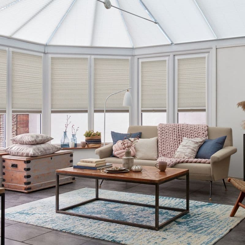 Cellular honeycomb pleated blinds lowered across tall conservatory windows, filtering soft daylight; beige sofa with chunky pink throw, wooden coffee table and blue patterned rug in a bright sitting area.