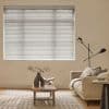 White horizontal blinds covering a window, closed and filtering daylight; beige living room with a low wooden coffee table, sculptural vase and open book, sofa and adjustable floor lamp.