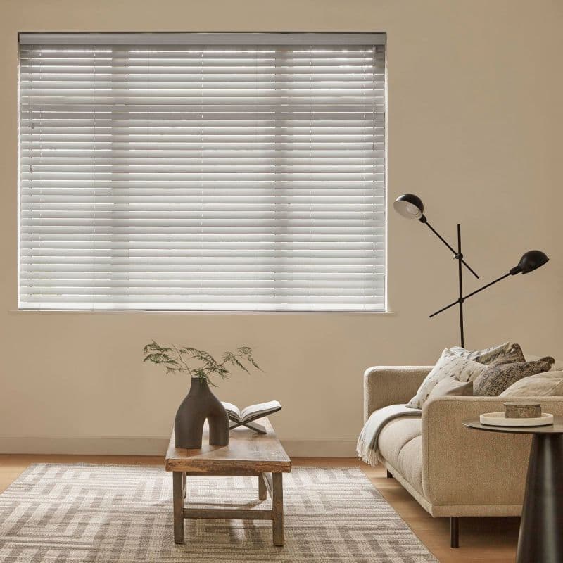 White horizontal blinds covering a window, closed and filtering daylight; beige living room with a low wooden coffee table, sculptural vase and open book, sofa and adjustable floor lamp.