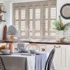 Beige louvered plantation shutters covering a wide kitchen window, slats partially tilted filtering soft daylight; framed by a sink and wooden countertop, dining table set, subway-tile backsplash and plant.