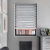 Grey zebra roller blind, lowered over window with alternating opaque and sheer horizontal stripes filtering light; grey kitchen wall with countertop espresso machine, radiator below and brick building visible outside.