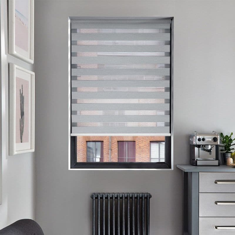 Grey zebra roller blind, lowered over window with alternating opaque and sheer horizontal stripes filtering light; grey kitchen wall with countertop espresso machine, radiator below and brick building visible outside.