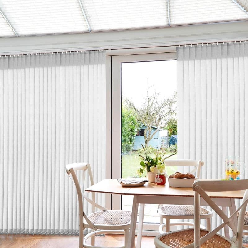 White vertical blinds covering glass doors, mostly closed with a central gap; filtering daylight and parted to reveal a backyard tree and shed beyond a dining table with chairs.