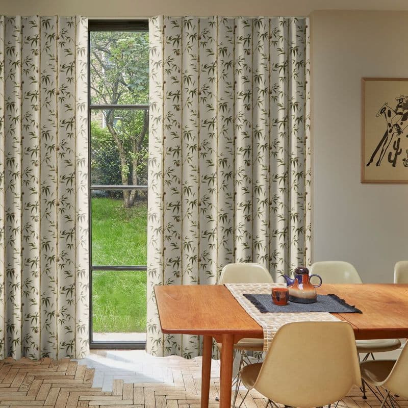 Floor-length cream curtains with green bamboo print, mostly drawn and slightly parted at a tall narrow window, filtering soft daylight into a dining room with wooden table and garden view.