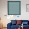 Teal pleated cellular blind, fully lowered with a narrow bottom reveal; filtering daylight and partially blocking a roof view — above a blue sofa in a modern, sunlit living room.
