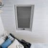 White pleated cellular blind in a roof skylight, fully lowered with horizontal folds, filtering daylight; set into white-painted tongue-and-groove ceiling above a bed with patterned cushions and a guitar cushion.