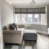 Gray fabric Roman blinds across a bay window, partially lowered and filtering daylight; living room with beige patterned corner sofa, gray ottoman, round side table, wood floor and street view.