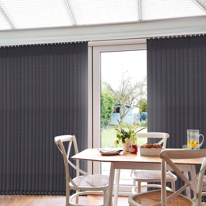 Dark grey vertical blinds, floor-to-ceiling and mostly closed, parted centrally to reveal a sunlit garden; a wooden dining table with chairs, vase of flowers, pastries and pitcher in foreground.