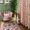 Full-length pink floral curtains at right, heavy fabric with botanical print; hung closed, softly filtering daylight; living-room corner with pink velvet chair, lamp, tall plant, shag rug, and cat.