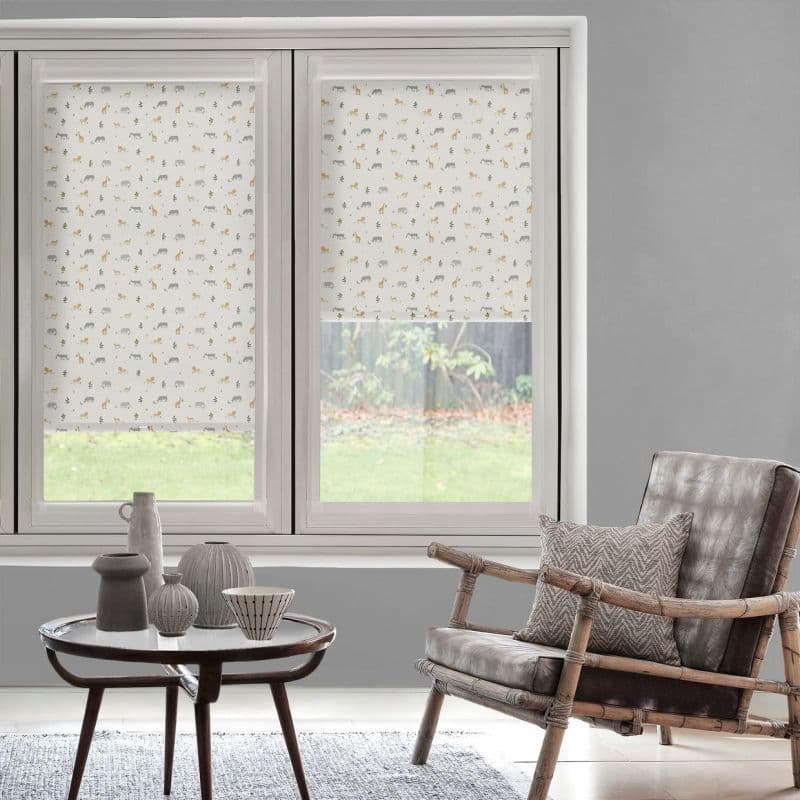 Printed roller blinds (white with small grey and ochre animal motifs) covering twin windows—left fully down, right half-raised—filtering daylight in a grey lit living room with rattan armchair and table.