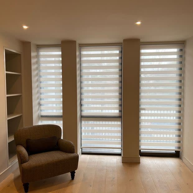 Zebra roller blinds covering three tall, narrow windows, with alternating opaque and sheer horizontal bands filtering soft daylight. Room: neutral living area with armchair, built-in shelves, recessed ceiling lights.