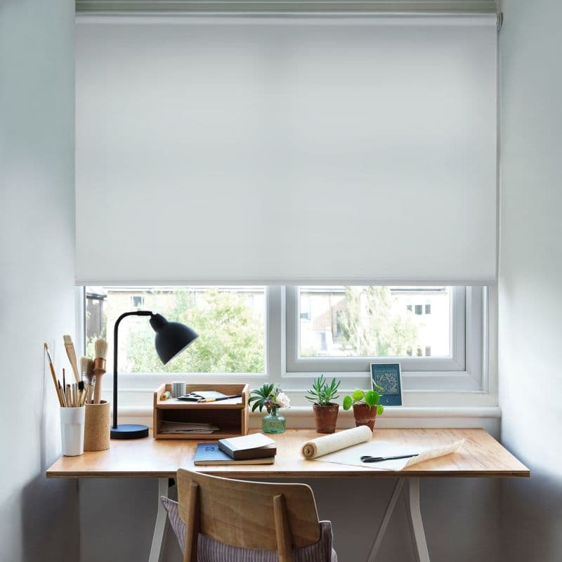 White roller blind, lowered across a double window with simple cassette, opaque and light-diffusing; filtering daylight. Context: tidy wooden desk beneath with lamp, brushes, notebooks and small potted plants.