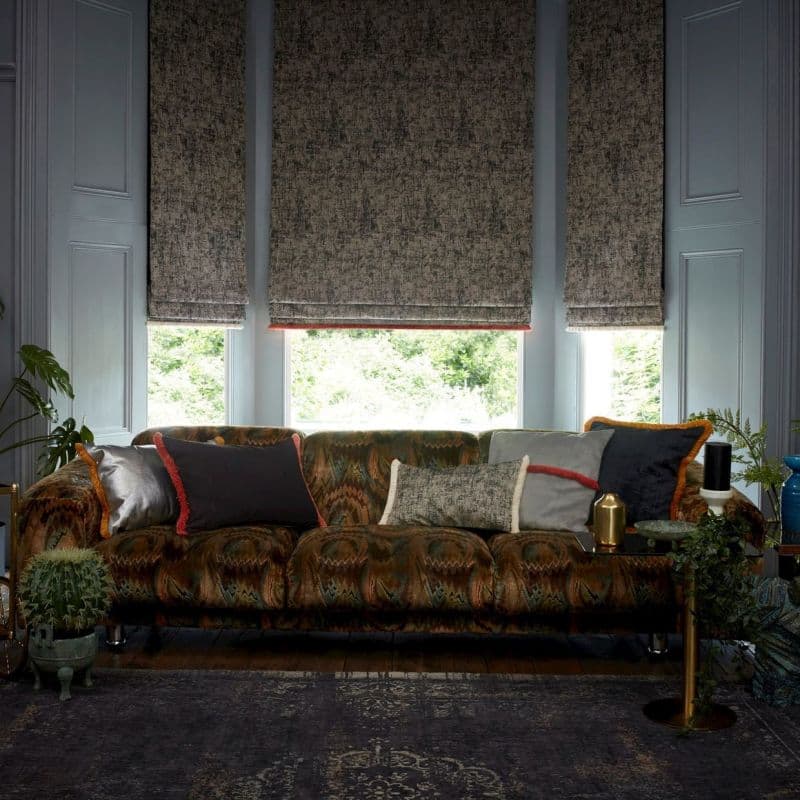 Three textured Roman blinds, lowered mid-height and filtering daylight; in front, a patterned tufted sofa with assorted cushions; indoor plants and side table in a softly lit living room.
