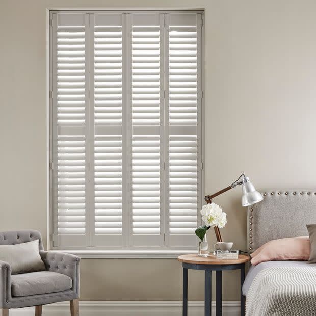 White full-height louvered plantation shutters covering a tall window, slats slightly tilted filtering soft daylight; bedroom setting with upholstered bed, bedside table and lamp, neutral walls and armchair.