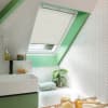 A white pleated skylight blind pulled down halfway, filtering soft daylight; set in a bright attic bathroom with green-painted frame, sloped ceiling, white herringbone tiles, sink, bathtub, and plant.