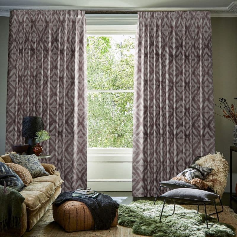 Full-length pleated patterned curtains with mauve-gray ikat motif frame a central sash window, parted slightly and filtering daylight; cozy living room with sofa, chair, textured rugs, leafy outside view.