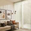 Pale fabric vertical blinds cover a floor‑to‑ceiling sliding glass door, slats angled to filter soft daylight, casting stripes; cozy living room with beige sofa, cushions, wall lamp and framed art.