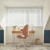 White horizontal blinds cover a wide window, slats tilted closed and filtering soft daylight; small attic study with a wooden desk, pink swivel chair, lamp, plant, and pale walls.