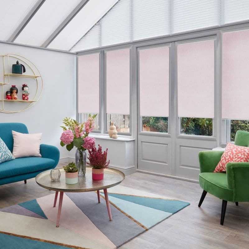 Pale pink roller blinds lowered over tall glass doors and windows, filtering daylight; bright conservatory-style living room with glass roof, teal sofa, green armchair, geometric rug, coffee table.