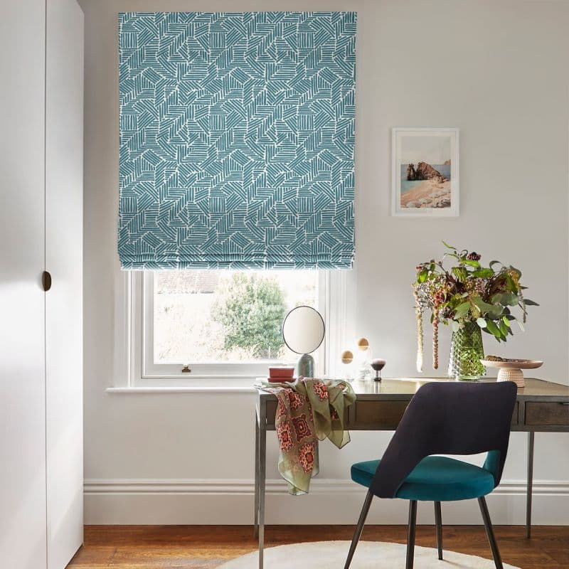 Teal geometric Roman blind, fully lowered, filtering soft daylight through a window; a wooden desk beneath holds a round mirror, vase of flowers, draped scarf, chair in a home office.