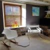 Wooden horizontal blinds covering a recessed window, lowered with slats tilted to filter sunlight; setting: cozy living room with stone sill, colorful glass vases, two modern chairs and soft carpeting.