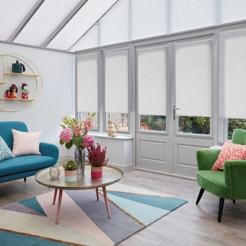 White roller blinds covering tall glazed French doors and sidelights, lowered to mid-height filtering daylight, in a bright conservatory-style living room with colorful sofas, coffee table, and garden view.
