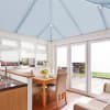 Blue pleated roof blinds fitted to the pitched conservatory ceiling, with white pleated window and door blinds lowered, filtering soft daylight over a dining area, kitchen sink, and garden view.
