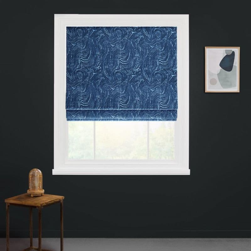 Blue patterned Roman blind, lowered and folded at bottom, with swirling texture; it blocks light over a white-framed window in a dark charcoal wall with side table and abstract art.