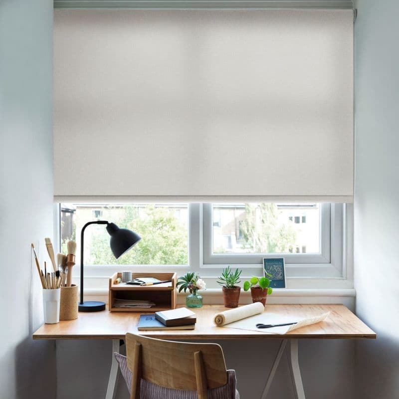 Beige roller blind, lowered nearly fully to cover the window, diffusing daylight; a wooden desk beneath holds a lamp, paintbrushes, notebooks, potted succulents and a chair in a home workspace.