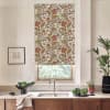 A floral-patterned roller blind covers a kitchen window above a stainless faucet, flanked by countertop plants, a framed sketch on the wall, and wooden cabinets beneath a marble countertop.