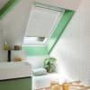 White pleated skylight blind, partially lowered over a slanted roof window, filtering daylight; set above a bathtub in a bright bathroom with green trim, white herringbone tiles, sink and plant.