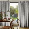 Floor‑length pleated gray patterned curtains, drawn aside, framing a glass door and filtering soft daylight into a dining nook with wooden table, pendant lamp and garden view.