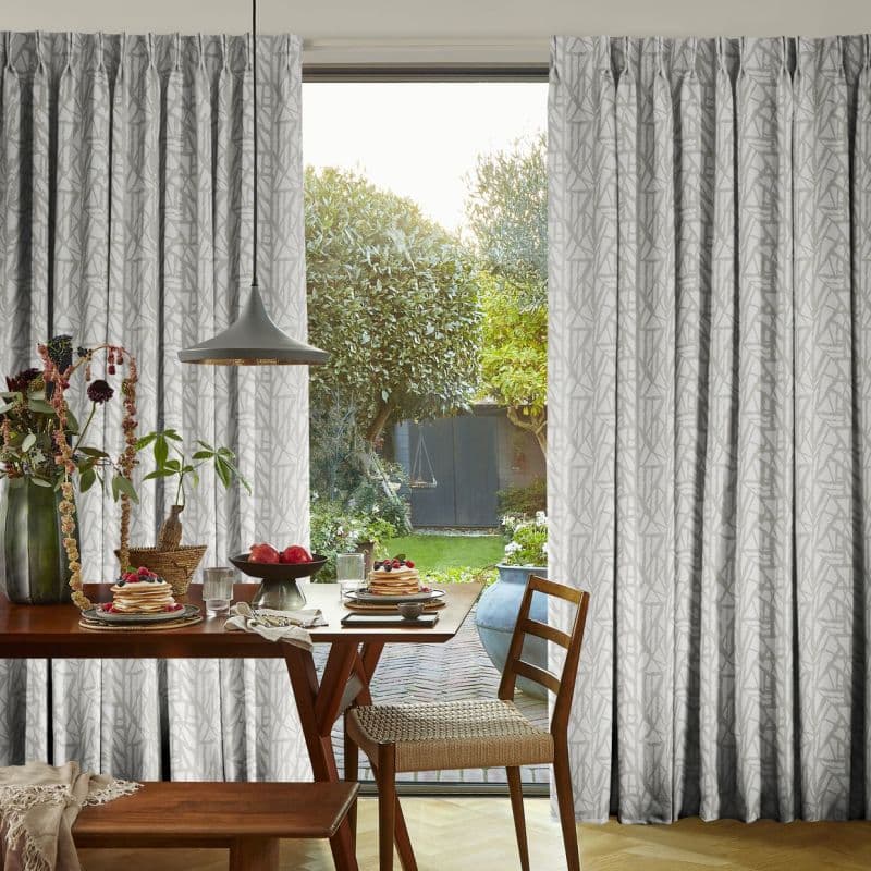 Floor‑length pleated gray patterned curtains, drawn aside, framing a glass door and filtering soft daylight into a dining nook with wooden table, pendant lamp and garden view.