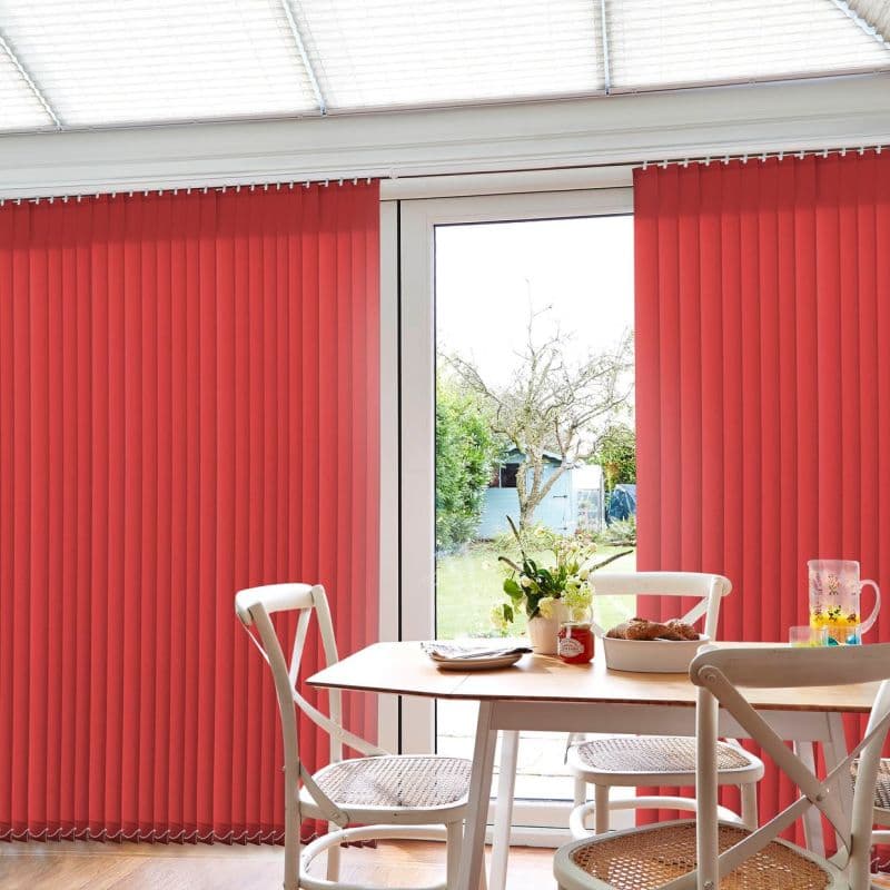 Red vertical blinds, full-height pleated slats across a sliding patio door, mostly closed with a central gap; sunlit dining table and white chairs framing a backyard lawn and shed.