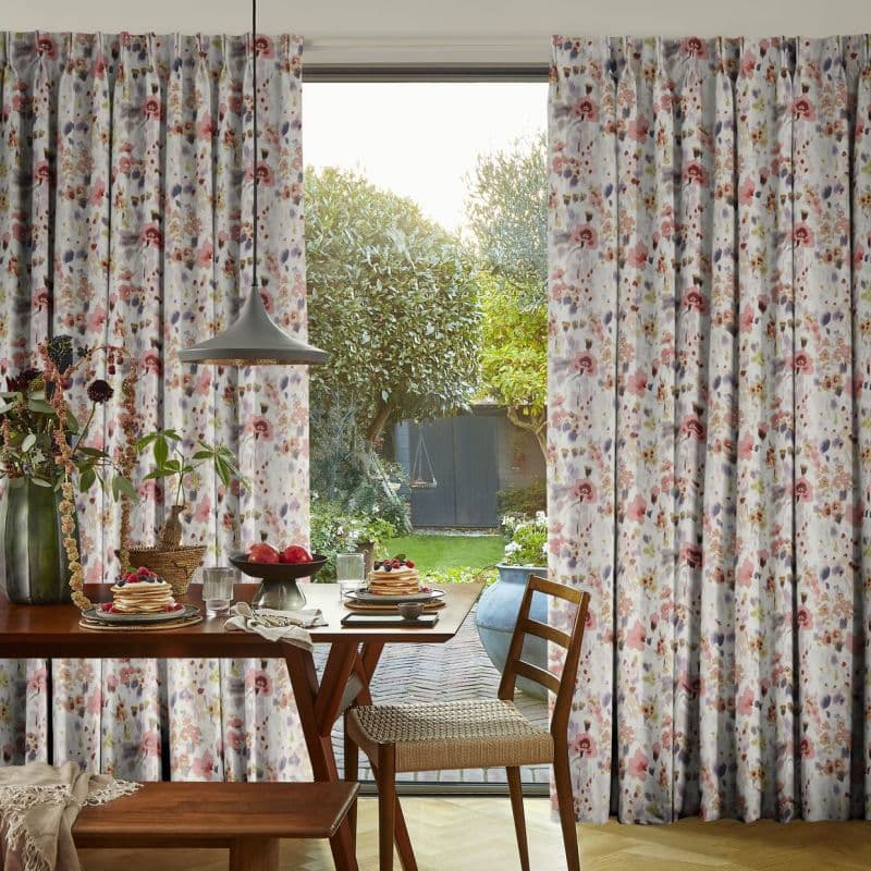 Floral pleated full-length curtains patterned with pink, mauve and grey blossoms frame a sliding glass door, partially drawn to filter light, overlooking a sunlit backyard garden and a breakfast table.