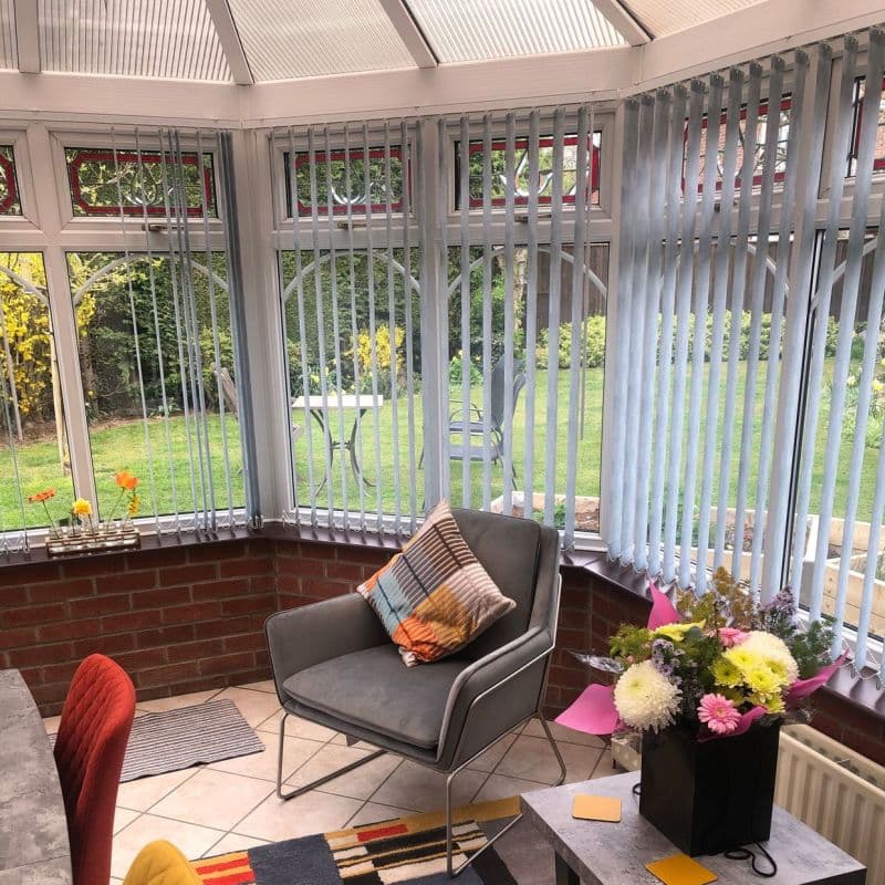 Pale-gray vertical blinds hang across wraparound conservatory windows, slats partially angled and filtering daylight; interior shows a tiled sunroom with an armchair, cushion, small table and garden view.