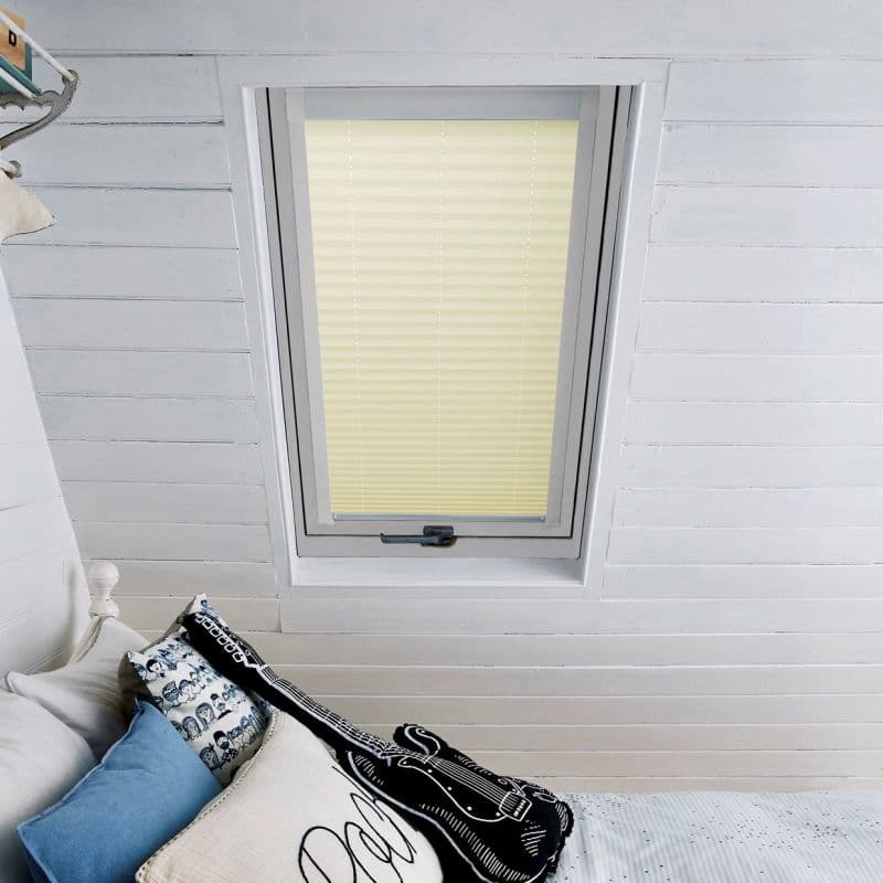 A pale yellow pleated blind, fully lowered over a skylight, diffuses soft daylight in a white-painted sloped attic ceiling above a bed adorned with pillows, including a guitar-shaped cushion.