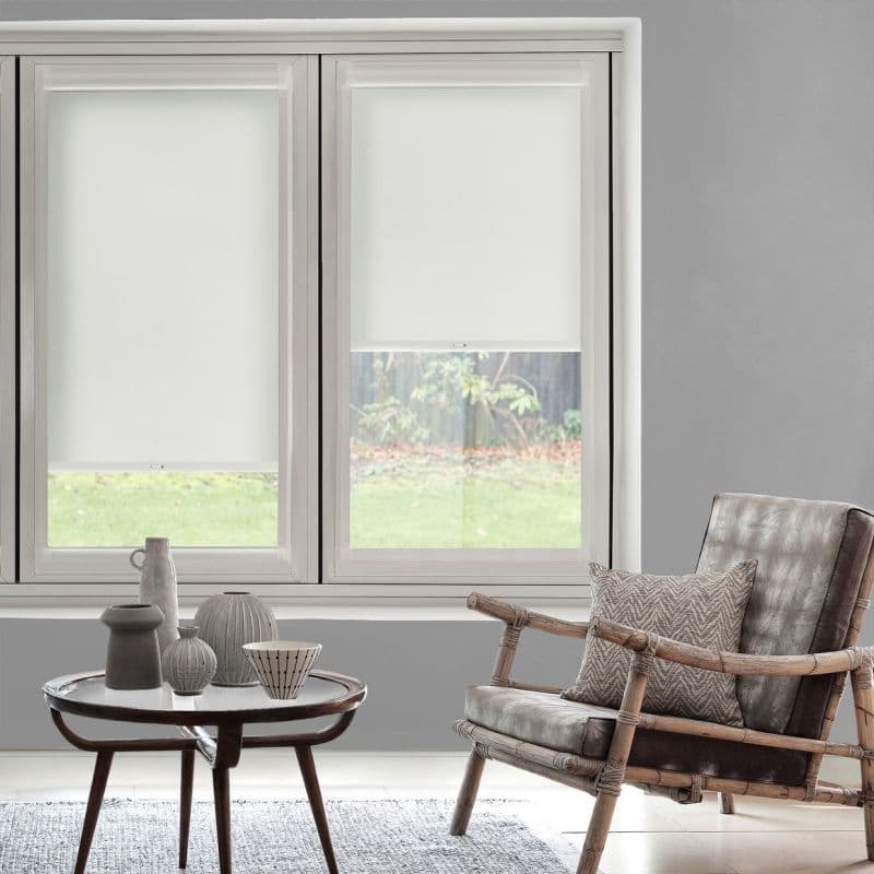 White roller blinds on two adjacent windows — left fully lowered, right halfway — filtering light over a lawn view; grey living area with a rattan armchair and small round coffee table.