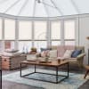Pleated cellular blinds, fully lowered on bay windows with light‑filtering opaque fabric and narrow horizontal pleats; filtering soft daylight into a conservatory‑style living room with sofa, coffee table, rug.