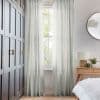 Sheer floor-length pale-gray curtains, pleated and hung at a rod, filtering soft daylight through a tall window; bedroom context with dark paneled wardrobe left, wooden nightstand and round mirror.