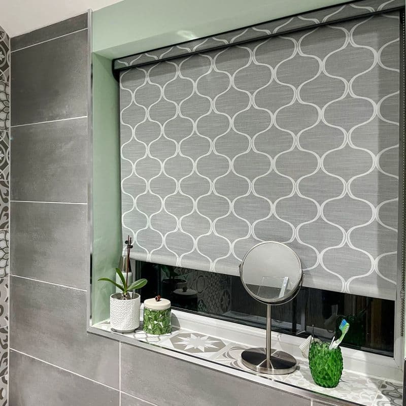 Grey patterned roller blind lowered, blocking light; sits over a bathroom window with tiled sill holding a small round makeup mirror, potted plant, jar, and toothbrush holder.