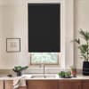Black roller blind covers most of a kitchen window, blocking light above a stainless-steel sink; surrounding countertop holds a bowl of greens, potted plants, a framed sketch, and wooden cabinets.