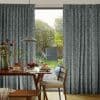 Floor-length grey patterned curtains, pleated and partially drawn to reveal a central glass door, filtering soft daylight, in a dining room with wooden table, pendant lamp and leafy garden beyond.