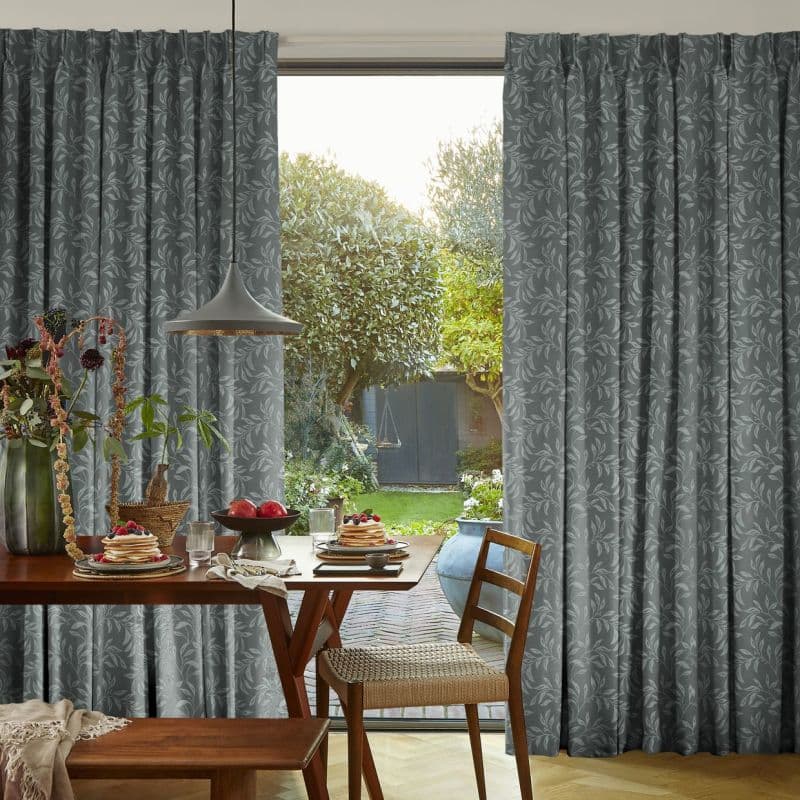 Floor-length grey patterned curtains, pleated and partially drawn to reveal a central glass door, filtering soft daylight, in a dining room with wooden table, pendant lamp and leafy garden beyond.
